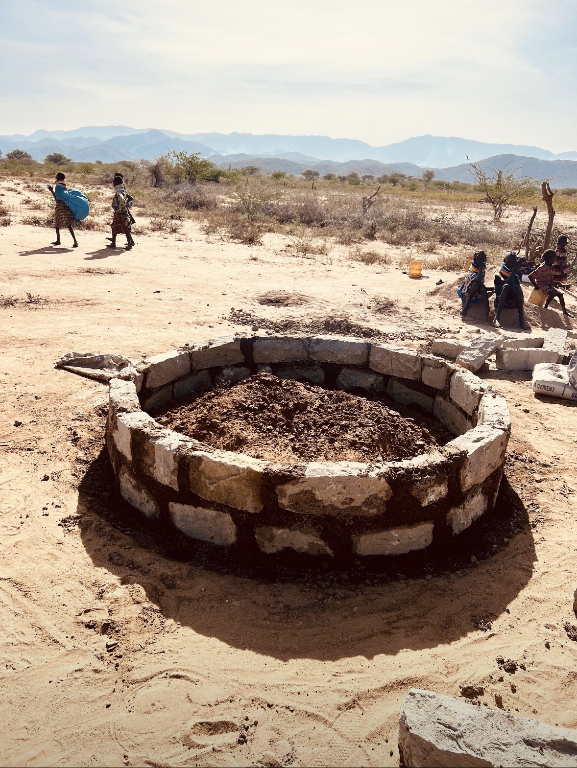 Stone well collar under construction in arid Turkana landscape with cement bags and children nearby